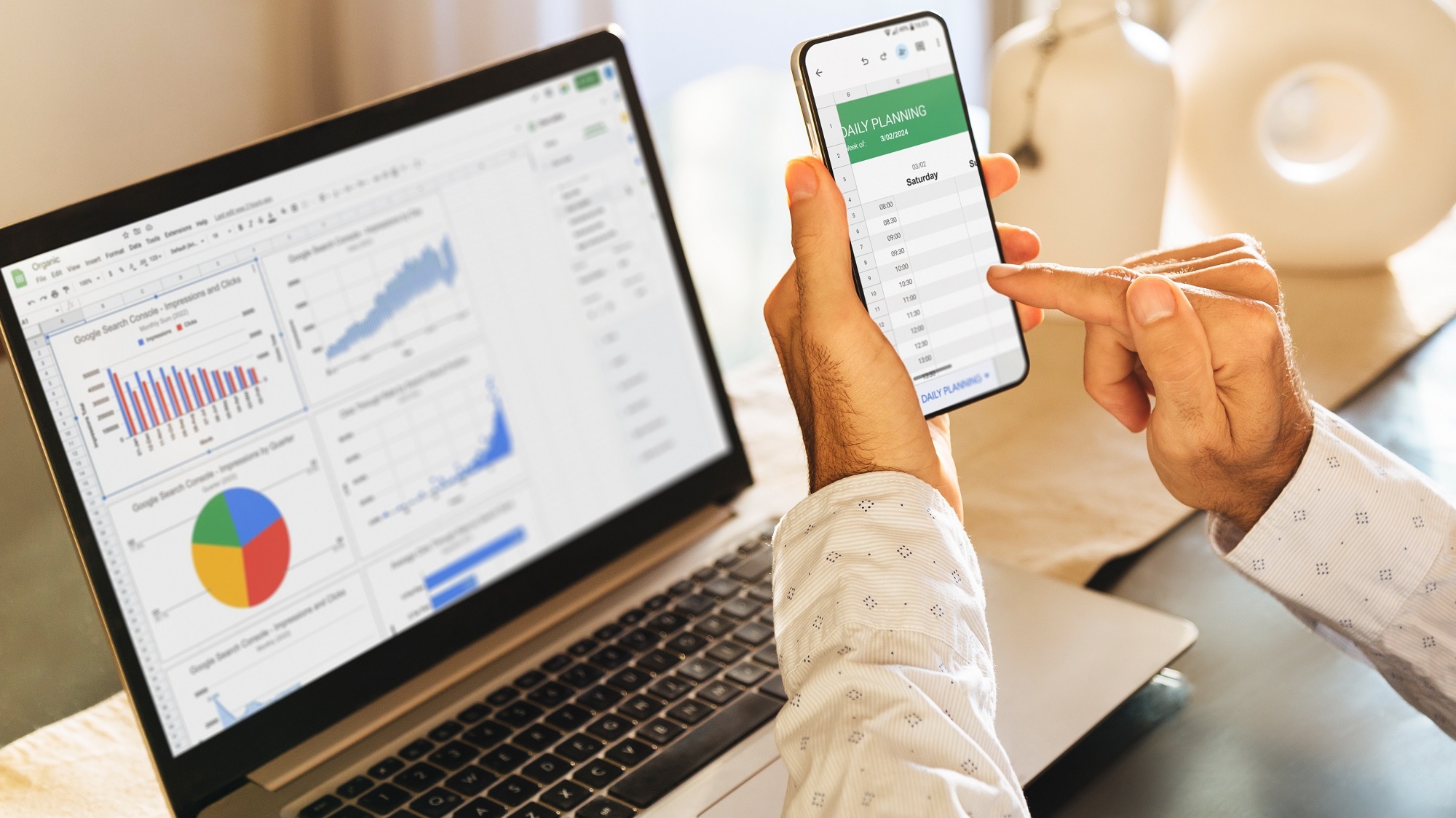 A stock photo of a person on their phone looking at a spreadsheet while several graphs are displayed on the laptop in front of them.